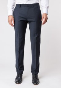 Tailored navyblaue Hose aus glattem Stoff, mit schmalem Schnitt, scharfen Falten und zwei Seitentaschen. Getragen mit schwarzen Schuhen.