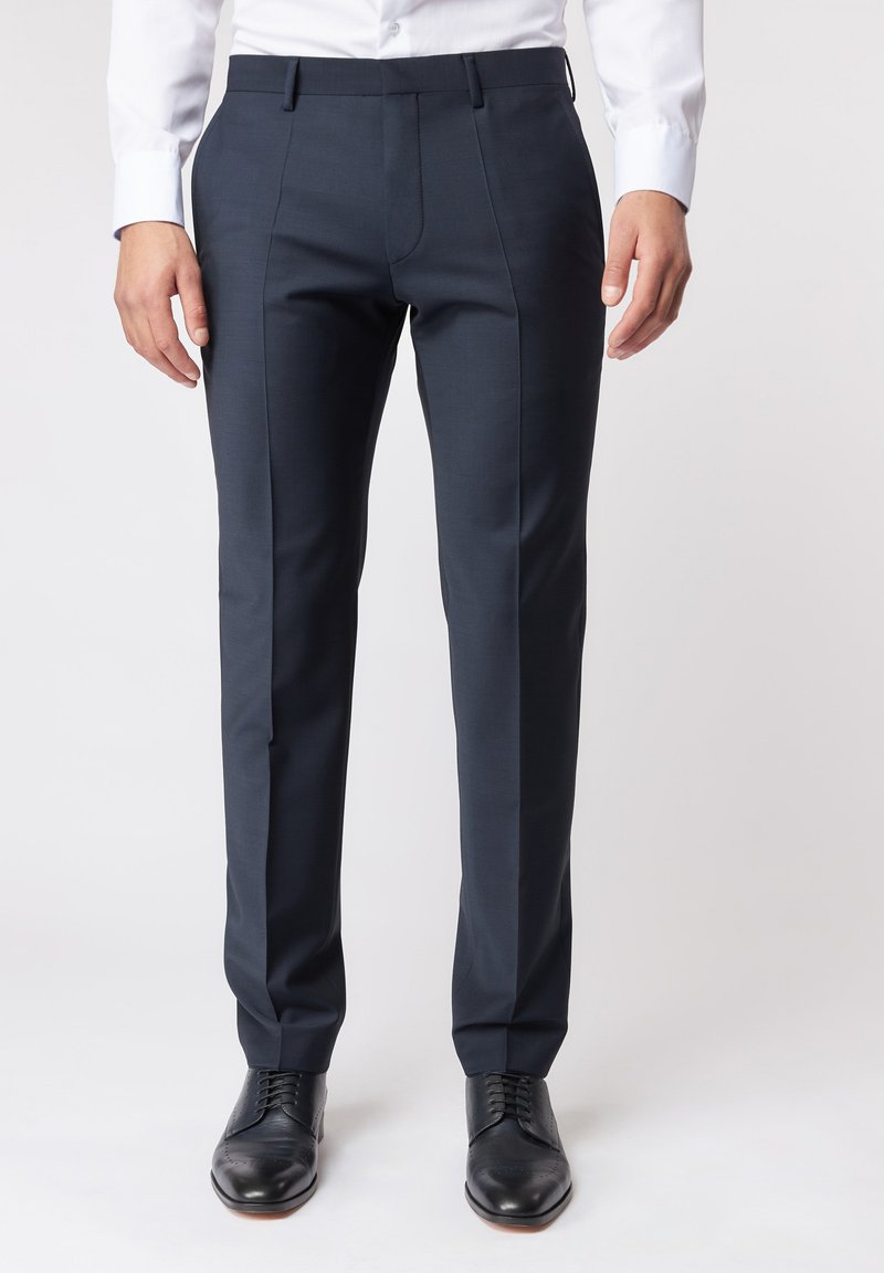 Tailored navyblaue Hose aus glattem Stoff, mit schmalem Schnitt, scharfen Falten und zwei Seitentaschen. Getragen mit schwarzen Schuhen.
