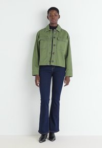 Olivengrønn cropped jakke med to brystlommer, svart turtleneck og mørkeblå slengjeans; modell iført svarte hæl støvler.