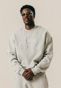 Młody czarnoskóry mężczyzna w okularach i beżowym swetrze z napisem "BLKVIS", stojący na tle jednolitego jasnego tła.