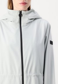 Light gray hooded jacket made from smooth, waterproof fabric featuring a front zipper and a black logo patch on the sleeve.