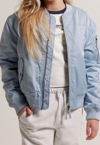 Giacca bomber azzurro chiaro con dettaglio trapuntato sul davanti, chiusura a zip, tasche laterali e polsini a costine, abbinata a una camicia leggera e pantaloni della tuta.