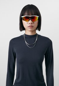 Lunettes de soleil avec une monture blanche courbée et des verres miroités jaunes-rouges. Portées avec une chemise à manches longues noire avec des accents texturés.