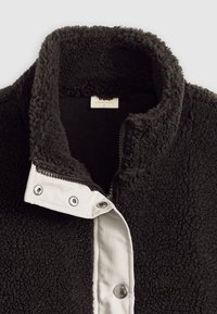 Chaqueta de forro polar negra con cuello alto, cierre de broche blanco y forro interior liso. Textura acogedora, con cremallera y herrajes plateados.