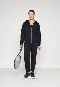 Νεαρή γυναίκα σε μαύρο φούτερ με φερμουάρ και αθλητικές φόρμες κρατά ένα ρακέτα τένις, στέκεται μπροστά από έναν απλό φωτεινό φόντο.