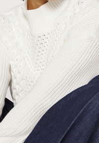 Pull blanc en maille avec texture côtelée, motifs torsadés et col montant, associé à un tissu bleu foncé.