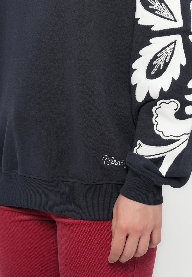 Zwarte sweatshirt met witte bloemenborduursel op de mouw; heeft een geribbelde onderkant en manchetten. "Wrong" is geborduurd nabij de onderkant.