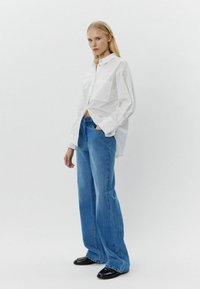 Maglietta bianca a maniche lunghe con una singola tasca, abbinata a jeans a gamba larga blu chiaro e scarpe nere; tessuto morbido e vestibilità rilassata.