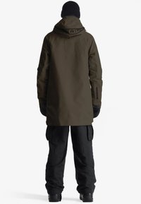 Giacca impermeabile khaki con cappuccio, dotata di polsini regolabili, abbinata a pantaloni cargo neri e stivali neri, mostrata da dietro.