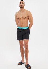 Zwarte zwembroek met een turquoise elastische tailleband en trekkoord. Glad textuur. Draag met zwarte schuif sandalen.