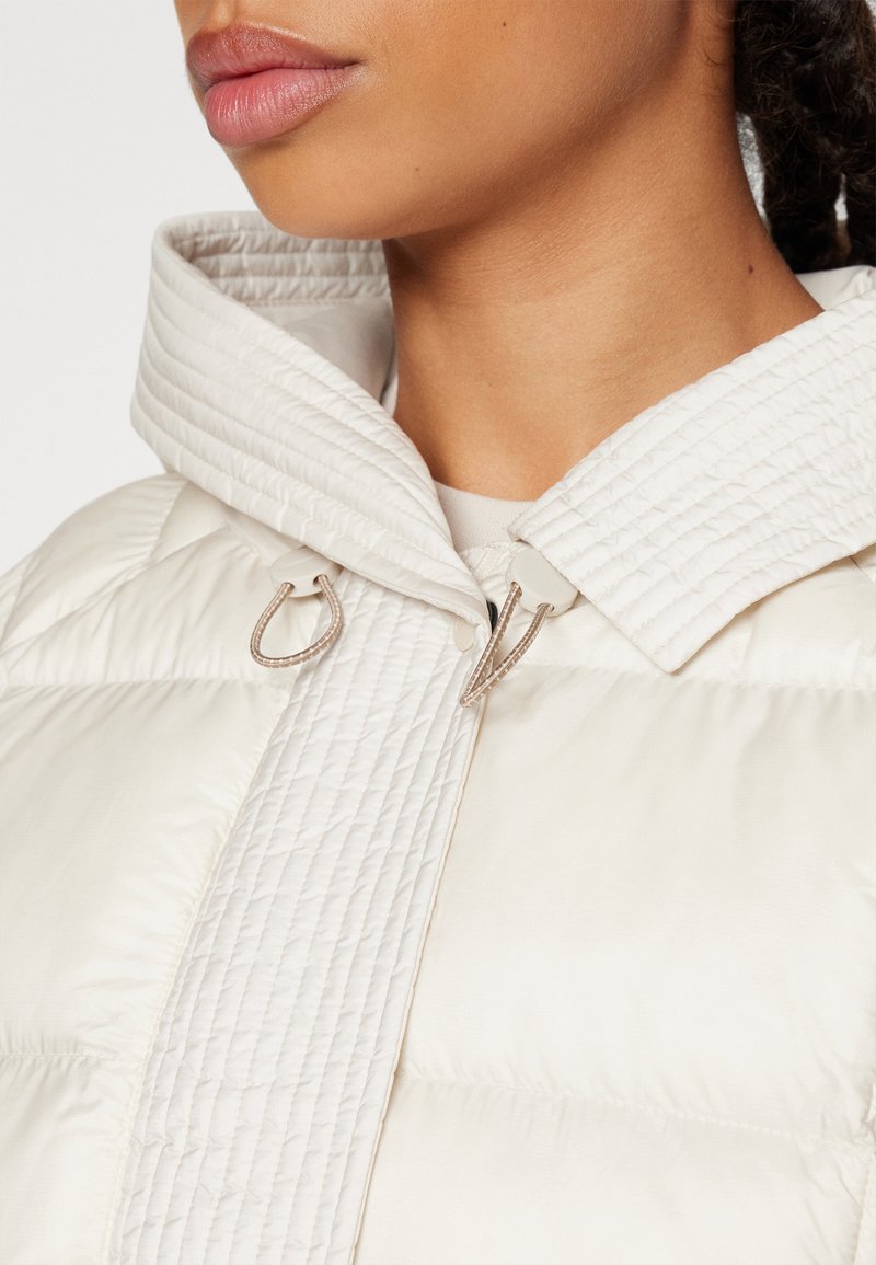 Giacca a vento di colore bianco sporco, con un design trapuntato, colletto alto e due chiusure a cordoncino al collo. Texture liscia e lucida.