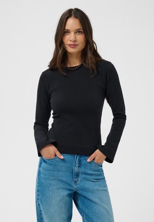 Femme aux longs cheveux bruns portant un t-shirt noir à manches longues et un jean bleu, debout les mains dans les poches devant un fond uni.