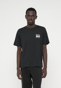 Μαύρο βαμβακερό μπλουζάκι με γραφικό σχέδιο σε λευκό χρώμα στην αριστερή πλευρά του στήθους, με άνετη εφαρμογή και κοντά μανίκια.