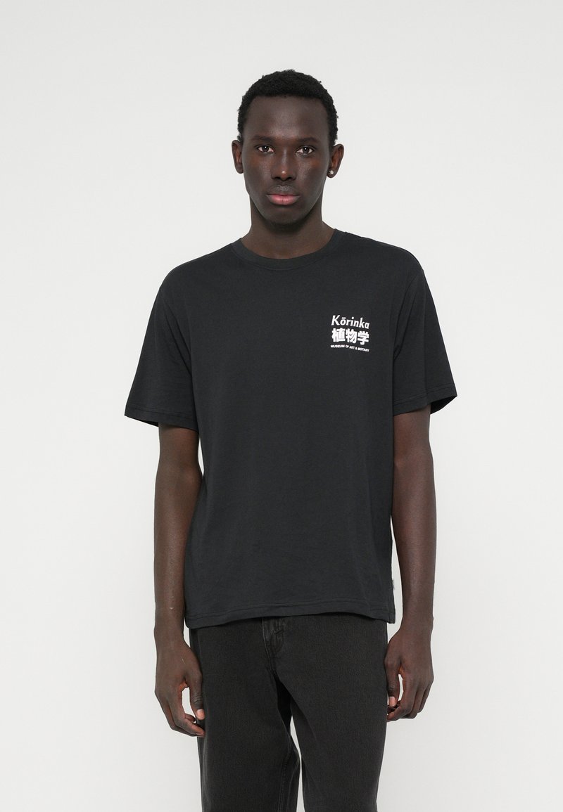 Μαύρο βαμβακερό μπλουζάκι με γραφικό σχέδιο σε λευκό χρώμα στην αριστερή πλευρά του στήθους, με άνετη εφαρμογή και κοντά μανίκια.