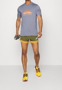 Hombre con camiseta azul de Nike Trail, pantalones cortos verdes y amarillos, y zapatillas de correr amarillas brillantes, sosteniendo una botella de agua gris, de pie sobre un fondo liso.