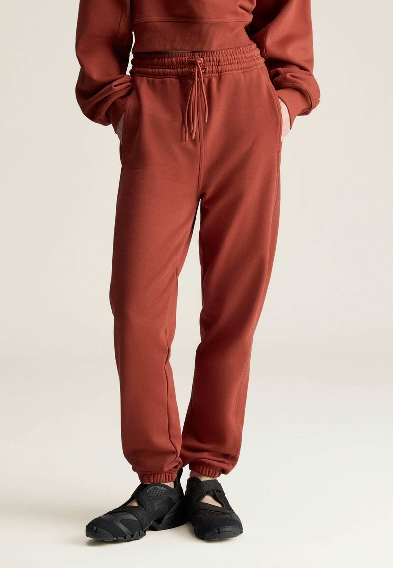 Osoba nosząca rdzawe spodnie dresowe z elastycznymi mankietami i sznurkiem w pasie, w połączeniu z czarnymi, przewiewnymi butami wsuwanymi.