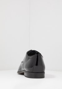 Scarpa in pelle verniciata nera con finitura liscia, punta rotonda e branding sottile. Presenta un tacco basso e un design classico.