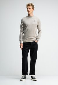 Jonge man die met zijn hand in zijn zak staat, gekleed in een beige, gestructureerde trui, zwarte opgerolde broek en zwarte hoge sneakers, voor een witte achtergrond.