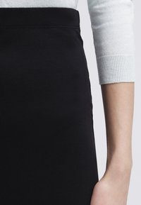 Gonna nera aderente con texture liscia e cintura elasticizzata. Manica di un maglione bianco parzialmente visibile, che mostra il dettaglio a costine del polsino.