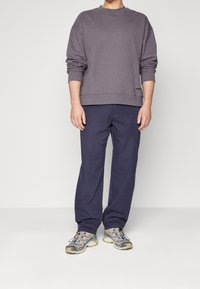 Un homme portant un sweat-shirt gris ample à manches longues, un pantalon droit bleu foncé et des chaussures de sport multicolores se tenant devant un fond uni.