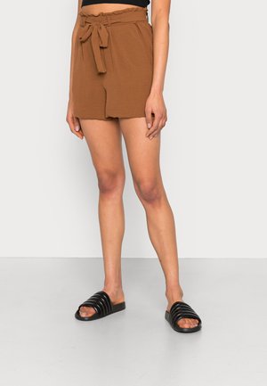 Frau trägt braune Shorts mit hoher Taille und einem Stoffgürtel, der an der Taille gebunden ist, sowie schwarze offene Sandalen auf einem grauen Boden.