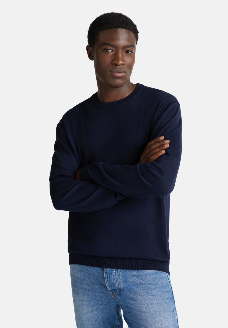 Marineblauer Strickpullover mit langen Ärmeln und Rundhalsausschnitt, kombiniert mit hellblauen Jeans. Einfaches Design, glatte Textur und lässige Passform.