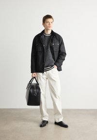 Black jacket with two chest pockets over a patterned sweater, paired with off-white pants and black shoes, holding a black leather bag.
