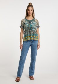 Blusa a fantasia con maniche corte e volants; i colori includono turchese, giallo e marrone; vita arricciata; indossata con jeans blu e sandali marroni.