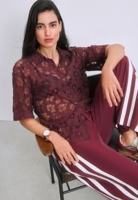 Jeune femme aux longs cheveux noirs porte une chemise en dentelle florale bordeaux, un pantalon bordeaux à rayures blanches, assise de manière décontractée sur une chaise.