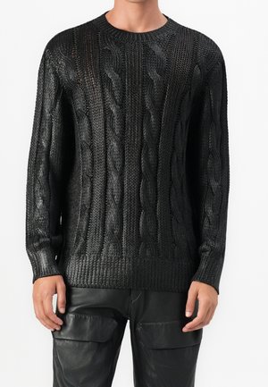 Homme portant un pull en maille torsadée noire et un pantalon en cuir noir, debout devant un fond blanc uni.
