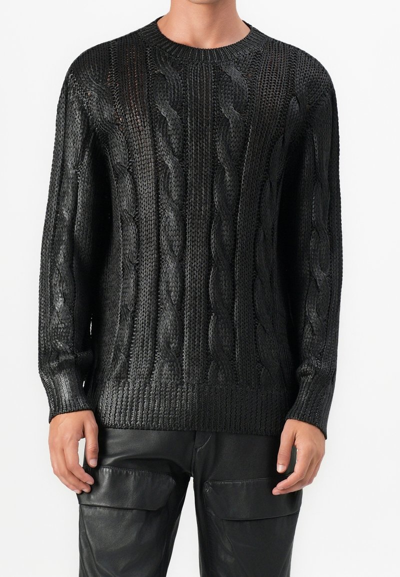 Homme portant un pull en maille torsadée noire et un pantalon en cuir noir, debout devant un fond blanc uni.