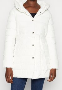 Chaqueta de plumas blanca con cintura cinturón, que cuenta con capucha, cierres de botones plateados y textura acolchada vertical. Material suave y ligero.