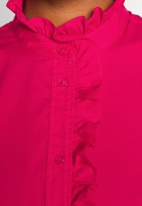 Magentová blůza s volánovým límcem, výraznými knoflíky a hladkou texturou látky. Úzce přizpůsobený střih se viditelnými švy.