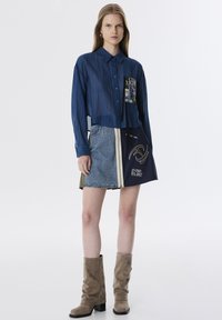 Denim overhemd in donkerblauw met verticale strepen, cropped ontwerp en een grafische patch, gecombineerd met een denim minirok en beige suède laarzen.