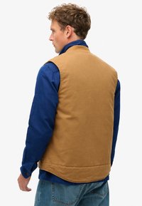 Gilet rembourré marron avec une texture lisse, présentant un col montant sur une chemise bleue à poignets boutonnés. Porté avec un jean en denim bleu.