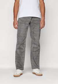 Šedé džíny mají uvolněný střih s širokými nohavicemi, viditelným šitím a dvěma předními kapsami. Jsou kombinovány s bílými teniskami.