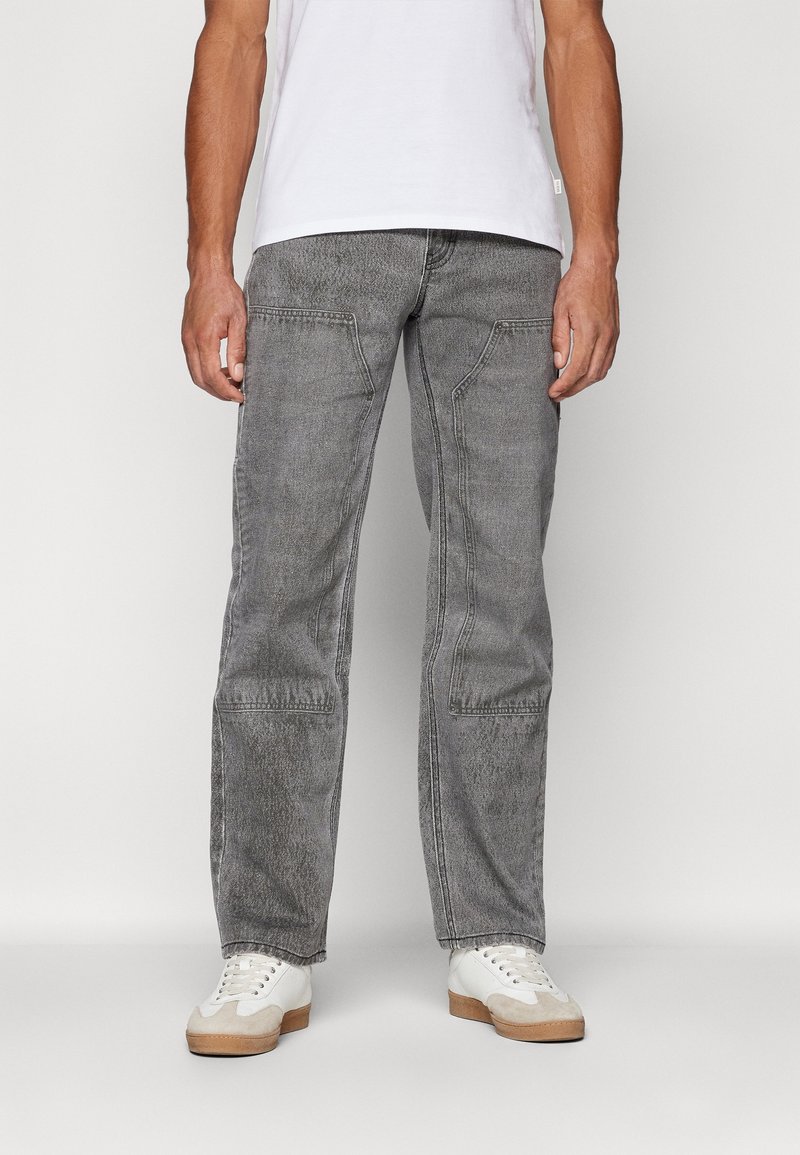 Šedé džíny mají uvolněný střih s širokými nohavicemi, viditelným šitím a dvěma předními kapsami. Jsou kombinovány s bílými teniskami.