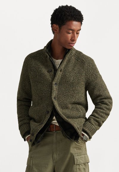 Cardigan en olive tricoté avec un col montant, une fermeture à boutons et un tissu texturé. Porté par-dessus une chemise légère en couches et un flanelle à carreaux.