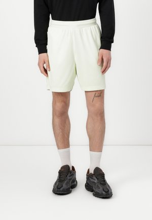 Shorts de sport vert clair en tissu doux, avec une taille élastique et des poches latérales. Portés avec des baskets noires et des chaussettes blanches.
