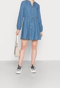 Denim dress with long puff sleeves, button closure, drawstring waist, and flared hem. Paired with a light beige handbag and white sneakers.