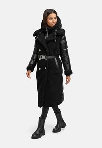 Lungo cappotto nero realizzato con un mix di materiali soffici e lucidi, design doppiopetto, vita con cintura e ampio cappuccio. Indossato con stivali neri.