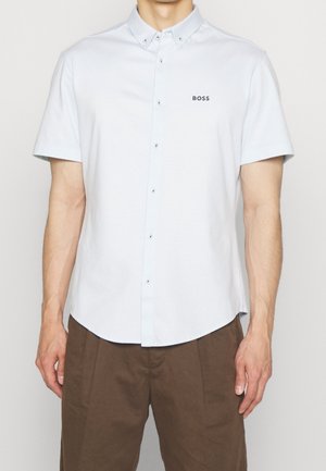 Mann trägt ein weißes kurzärmeliges Hemd mit dem "BOSS"-Logo auf der Brust und braune Hosen, steht vor einem einfarbigen Hintergrund.