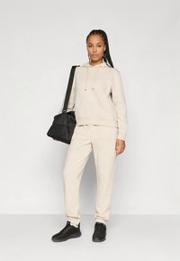 Γυναίκα με μπεζ φούτερ και φόρμες, μαύρα αθλητικά παπούτσια, κρατώντας μαύρη τσάντα ώμου, στέκεται μπροστά σε απλό ανοιχτό φόντο.