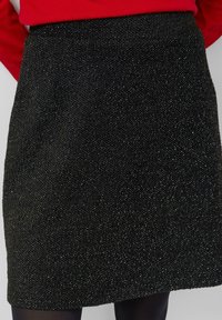 Schwarzer, strukturierter, knielanger Rock, getragen mit schwarzen Strumpfhosen und einem roten Pullover, von der Taille bis zur Mitte des Oberschenkels gezeigt.