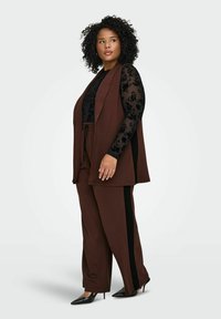 Hnědá vesta a odpovídající kalhoty s černými pruhy po stranách. Na vrchu je průsvitná černá květinová košile s dlouhými rukávy. Look doplňují lodičky na podpatku.