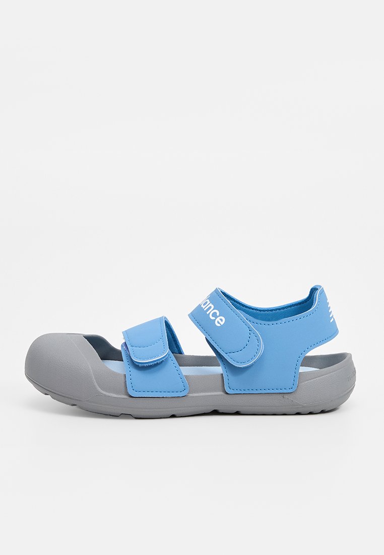 Sandalias de color azul claro con suelas de goma gris. Cuentan con correas ajustables de velcro y una puntera redondeada, diseñadas para la comodidad y la accesibilidad.
