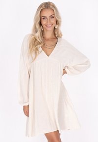 Rochie de culoare crem, cu decolteu în V, mâneci lungi tip bufant, țesătură texturată, modele în dungi verticale și accente metalice subtile.