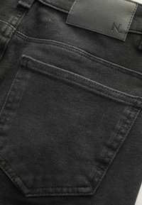 Zbliżenie na tylną kieszeń czarnych dżinsów z denimu z przeszyciami oraz czarną skórzaną naszywką z tłoczonym literą "N".