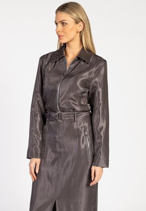 Frau mit langen blonden Haaren, die ein schimmerndes, dunkelgraues, mit einem Gürtel versehenes Kleid mit langen Ärmeln und einem Reißverschluss auf der Vorderseite trägt und zur Seite schaut.
