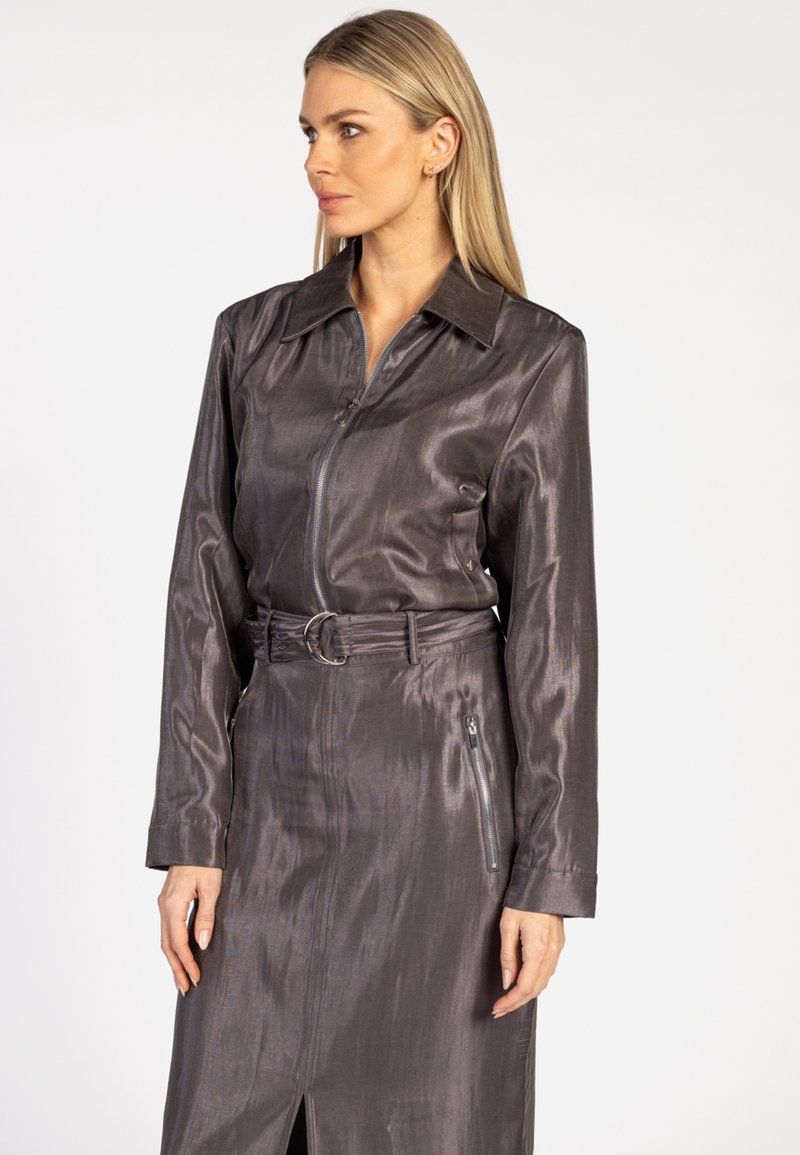 Vrouw met lang blond haar, gekleed in een glanzend donkergrijs jurkje met een riem, lange mouwen en een rits aan de voorkant, kijkend opzij.